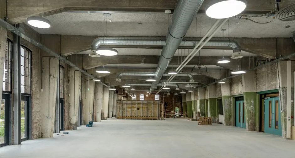 A vast empty warehouse with concrete walls and ceiling, metal ductwork, and large windows.